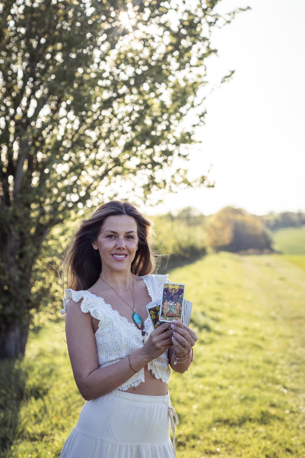 Eva Nunn standing in a sunlit field holding a tarot deck with The Lovers card